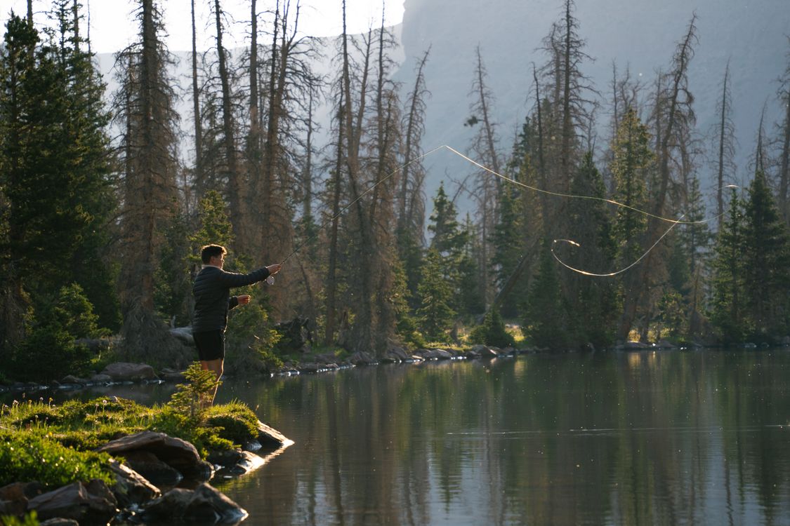 A person casting a fly-fishing line beside a forested alpine lake.