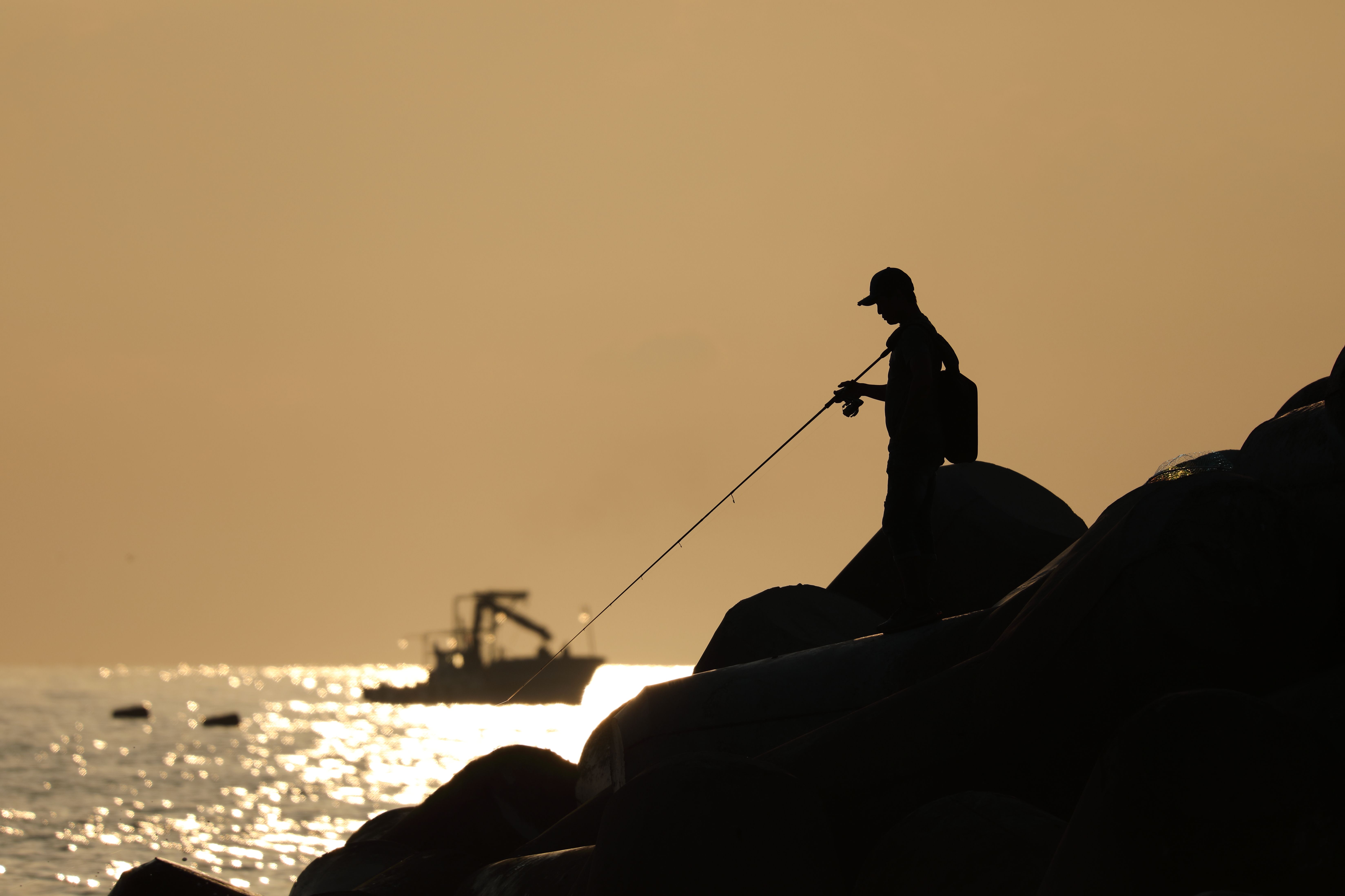 A person fishing with a rod and reel from coastal rocks beside the sea at sunset.