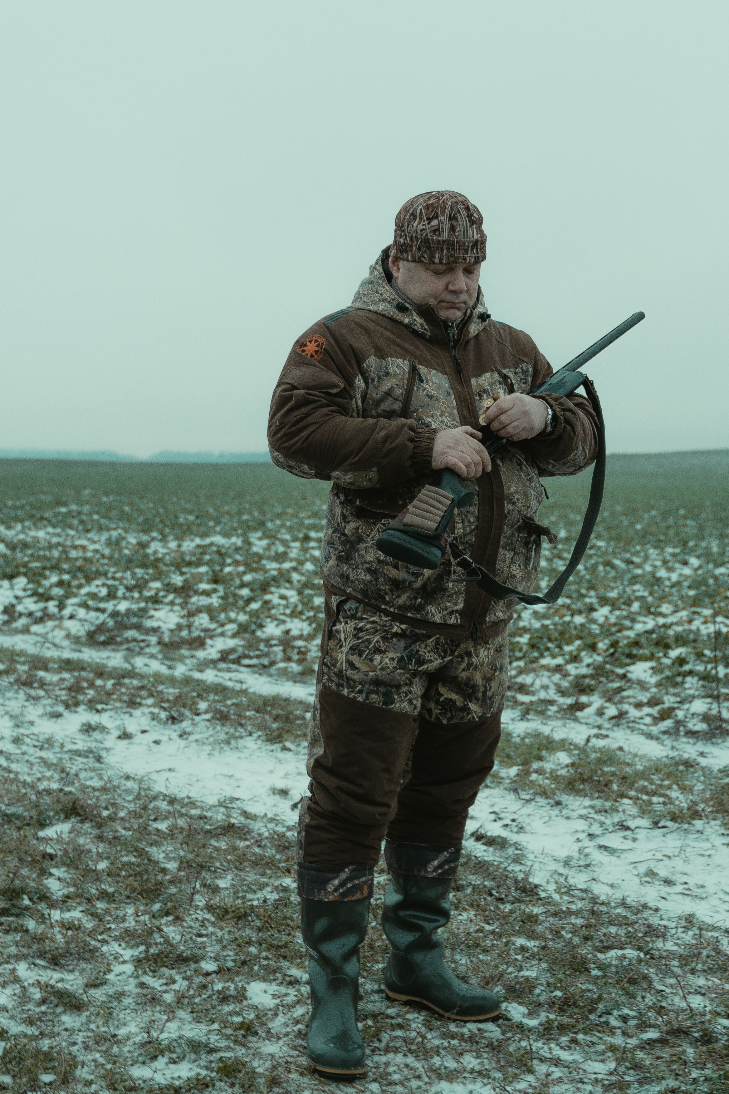 An adult hunter in camouflage standing in a snowy field with a hunting firearm.
