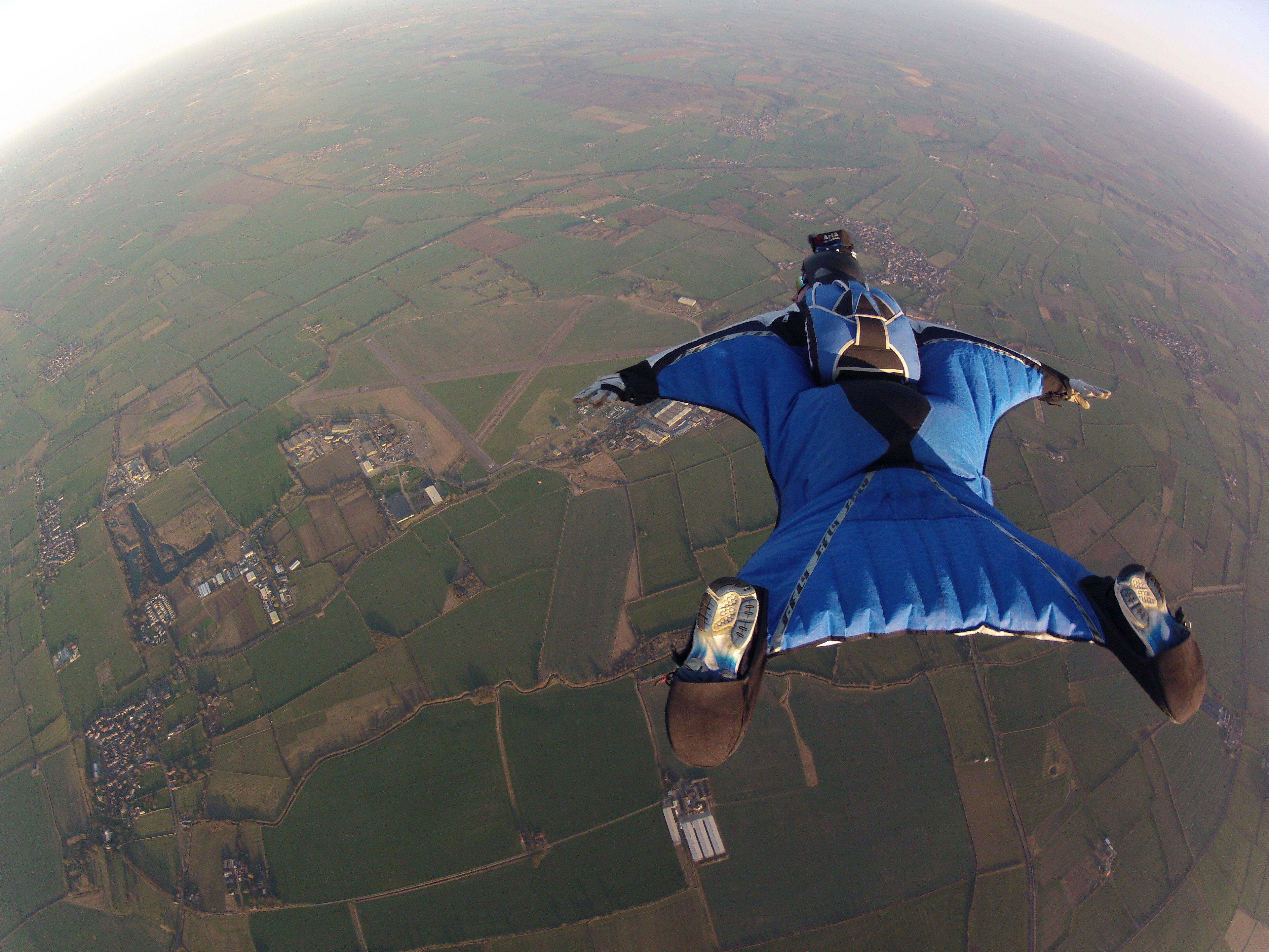 A person in a blue wingsuit flying above fields near Langar Airfield in the United Kingdom.
